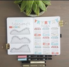 an open planner sitting on top of a table next to some pens and a potted plant