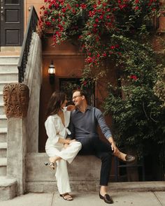 a man and woman sitting on the steps in front of a building with red flowers