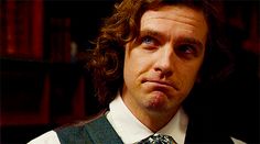 a man with long curly hair wearing a vest and tie in front of bookshelves