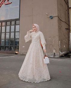 a woman in a white dress is standing on the street with a handbag and wearing a hijab