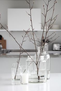 three glass vases with branches in them sitting on a counter top next to a candle