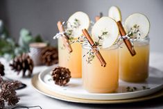 three glasses filled with apple cider sitting on top of a white plate next to pine cones