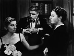 two women and a man standing in front of a bookshelf talking to each other