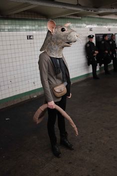 a person wearing a rat mask and holding a snake in a subway station with other people standing around