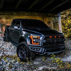 a black truck parked under a bridge in the woods with rocks and grass around it