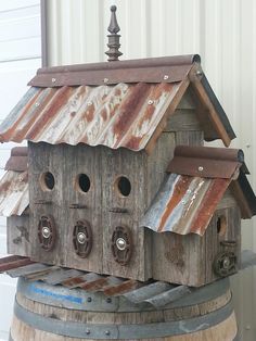 a wooden birdhouse sitting on top of a barrel next to a white building with rusted metal roof