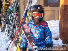 a young person holding skis and snowboards in front of a building with snow on the ground