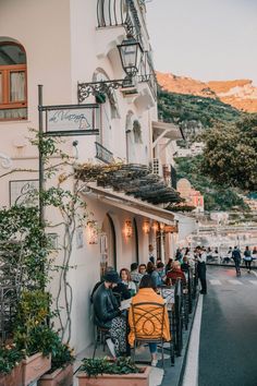 people sitting at tables in front of a building on the side of a road with mountains in the background