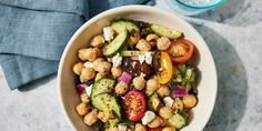 a white bowl filled with chickpeas, cucumbers and tomatoes next to a glass of water