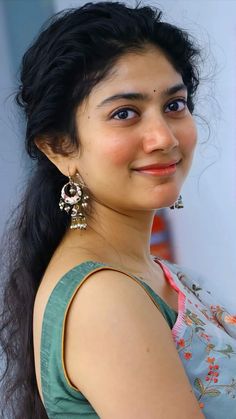 a woman with long black hair and earrings on her head smiles at the camera while standing in front of a white wall