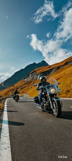 two motorcycles are driving down the road in front of some mountains and grass on either side