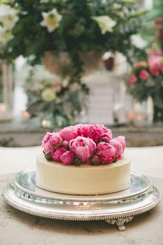 a white cake with pink flowers on top sitting on a silver platter in front of potted plants