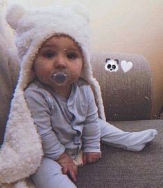 a baby sitting on a couch wearing a teddy bear hat with a pacifier in it's mouth
