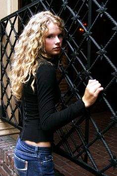 a beautiful young blonde woman standing next to a black iron fence with her hands in her pockets