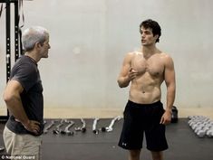 three men standing in a gym with no shirts on and one man without a shirt is looking at his cell phone