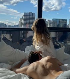 a man and woman laying in bed looking out the window at the city below them