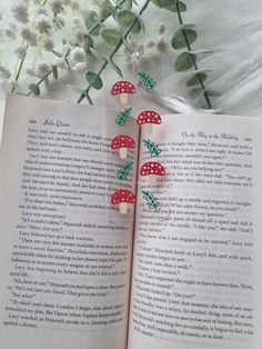 an open book with red mushrooms and green leaves on it next to some white flowers