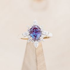 a ring with a purple stone surrounded by white diamonds on top of a wooden stand