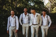 a group of men standing next to each other wearing white pants and suspenders, with trees in the background