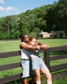 two people leaning on a fence in front of a field with trees and houses behind them