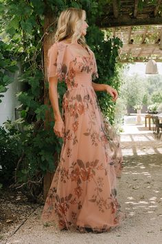 a woman in a long dress standing under a pergolian covered arbor with greenery