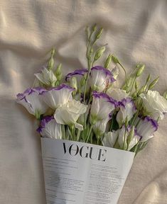 a bouquet of white and purple flowers in a paper vase on a bed with sheets