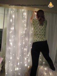 a woman standing on top of a bed in front of a window covered with white lights