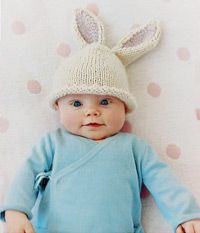 a baby wearing a knitted bunny hat and blue bodysuit laying on a polka dot blanket