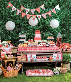 an outdoor picnic is set up with red and white plaid table cloths, paper lanterns, fruit baskets, and bunting banners