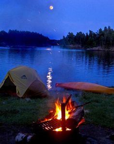 two tents set up next to a campfire on the shore of a lake at night