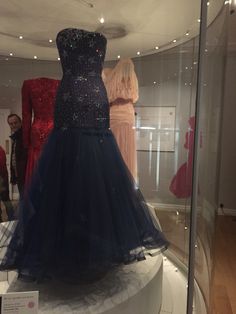 an evening gown on display in a glass case with other dresses behind it and people standing around