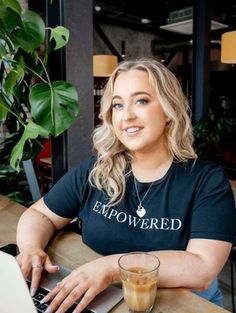 A woman with long blonde hair, wearing a black "EMPOWERED" t-shirt, sits at a table with a laptop and a glass of coffee in a cafe-like setting, smiling at the camera. With a lush indoor plant in the background, she perfectly captures the essence of being an empowered brand photographer. Outdoor Brand, Balloon Pop, What Image, Photographer Branding, Branding Photoshoot