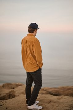 a man standing on top of a rock next to the ocean wearing a baseball cap