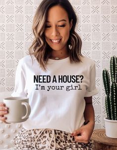 a woman holding a coffee mug and wearing a t - shirt that says need a house? i'm your girl