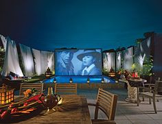 an outdoor movie is being projected on the wall behind wooden tables and chairs, while people sit around