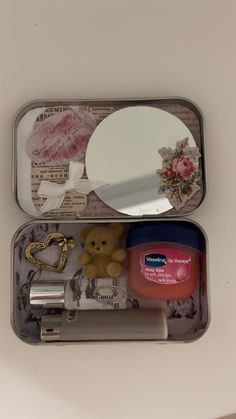 an open tin with a teddy bear, mirror and other items in it on a table