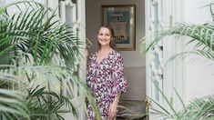 a woman standing in an open doorway with palm trees and potted plants behind her