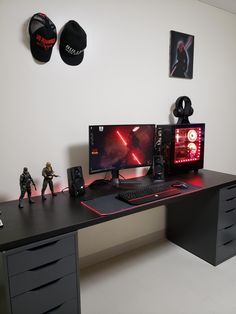 a desk with two computer monitors and toy figures on top of it, in front of a white wall