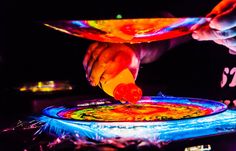 a person is painting on a plate with bright colors in the dark, while another hand reaches out to touch it