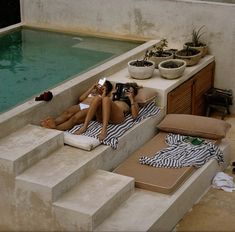 two women lounging in front of a swimming pool with towels on the floor and one woman taking a selfie