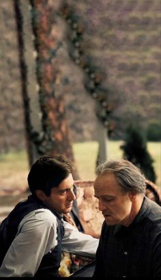 two men sitting next to each other in front of a tree and grass covered field