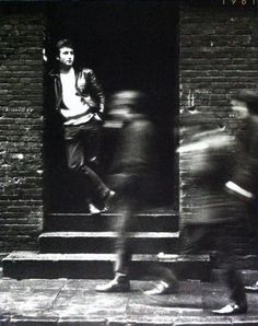 two men walking past a brick building with graffiti on the door and one man sitting down