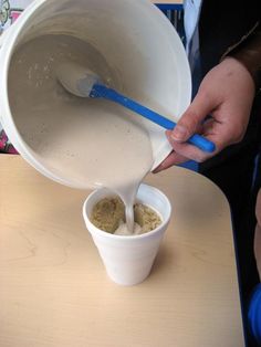 a person pouring something into a cup on top of a wooden table next to a blue plastic spoon