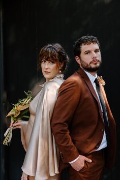 a man and woman standing next to each other in front of a black wall holding flowers