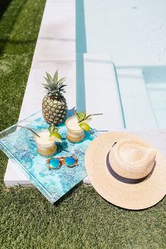 two pineapples are sitting on a towel next to some glasses and a straw hat