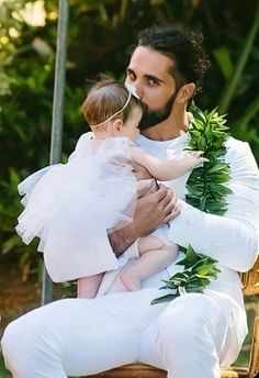a man holding a baby in his arms while sitting on a chair with greenery around him
