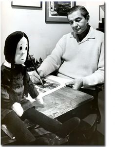 a man sitting at a table with a doll next to him and an adult holding a pen