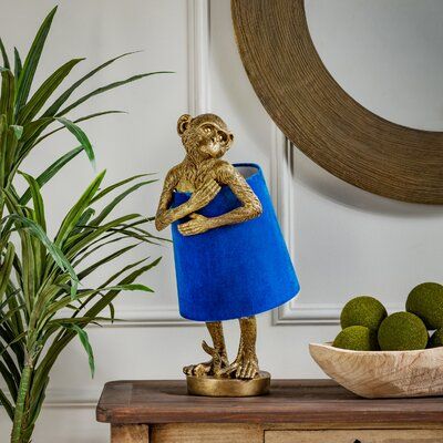 Funky monkey. This novelty lamp illuminates your space with an eclectic, maximalist vibe. It measures nearly 2' tall, and it's made from resin in a burnished gold hue and has a simple, circular base. The design features the shape of a cheeky monkey holding the lampshade around its middle like it's wearing a dress. We love that the class fabric shade comes in a luxe blue velvet for a pop of bright color. This table lamp plugs in with a standard cord and turns on with an in-line switch, and it tak Gold Monkey, Resin Fabric, Desk Lamps Bedroom, Pineapple Lamp, Novelty Lamps, Blue Table Lamp, Globe Lamps, Gold Table Lamp, Blue Table