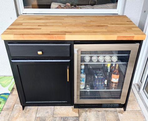 Outdoor bar with wooden countertop, black cabinet and drawer with gold handles on the left and stainless steel glass front mini-fridge on the right Outdoor Bar Fridge Ideas, Outdoor Bar With Mini Fridge, Coffee Bar Mini Fridge Cabinet, Diy Outdoor Bar With Mini Fridge, Cabinet Around Mini Fridge, Outdoor Bar With Refrigerator, Mini Fridge Cabinet Outdoor, Diy Bar Fridge Cabinet, Outdoor Fridge Cabinet Diy Patio