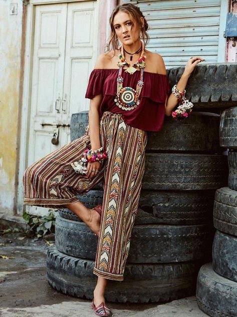 off-shoulder-wine-red-top-with-large-frill-combined-with-multicolored-trousers-in-tribal-print-boho-clothing-worn-by-brunette-woman-with-lots-of-chunk-jewelry-leaning-on-a-pile-of-truck-tires Brunette Jewelry, Bohemian Attire, Stile Pin Up, Bohemian Outfits, Outfits Boho, Stile Boho Chic, Boho Styl, Look Boho Chic, Estilo Hippy